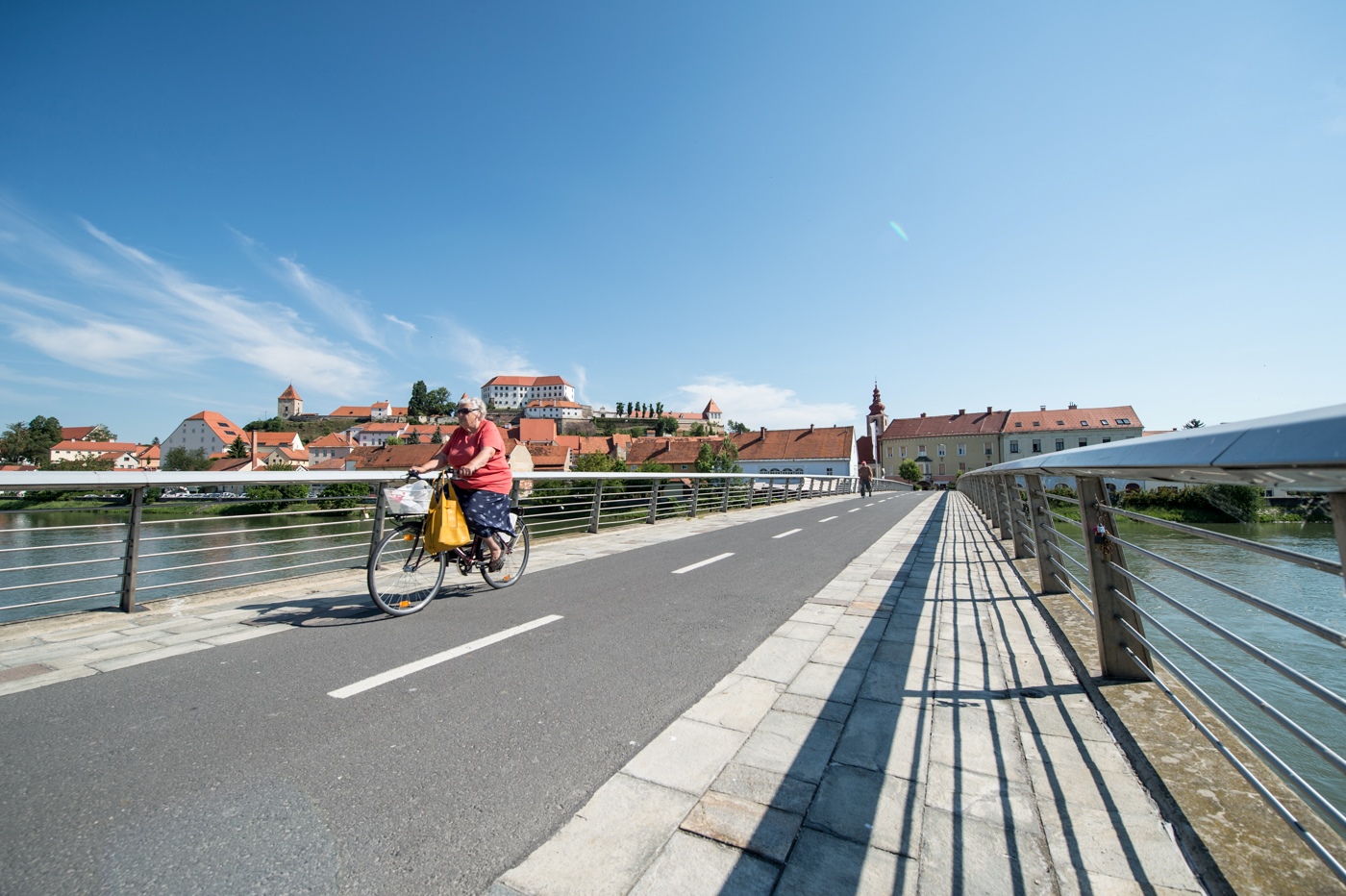 Starejša gospa na kolesu, kolesari po kolesarski stezi čez most, v ozadju vidno mesto Ptuj.